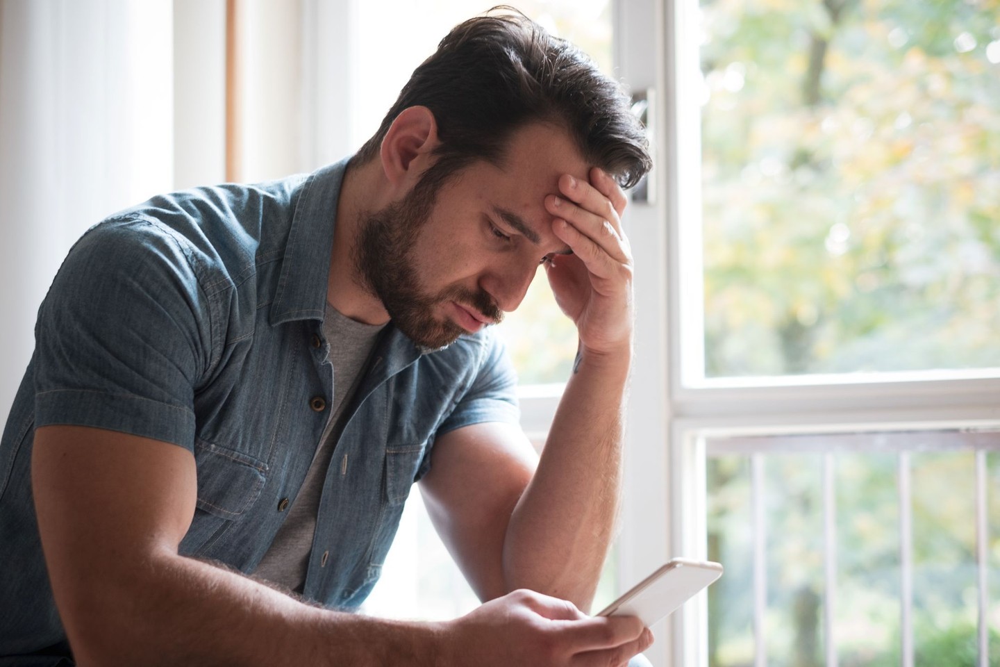 Worried man holding a smartphone with his hand on his forehead beside a window