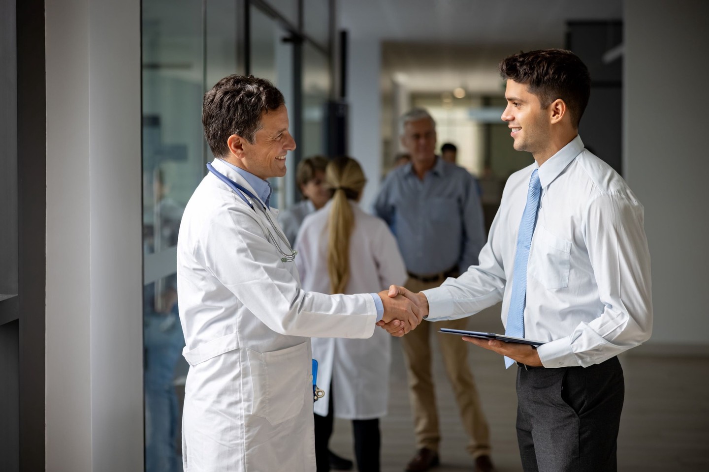 Doctor shakes hands with a businessman holding a tablet in a hospital corridor.