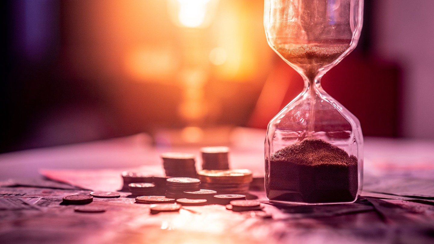 An hourglass set next to messy stacks of coins and cash bills, with a bright light in the background.
