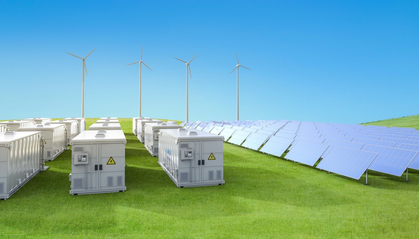 Rows of battery storage containers beside solar panels and wind turbines in a green field.