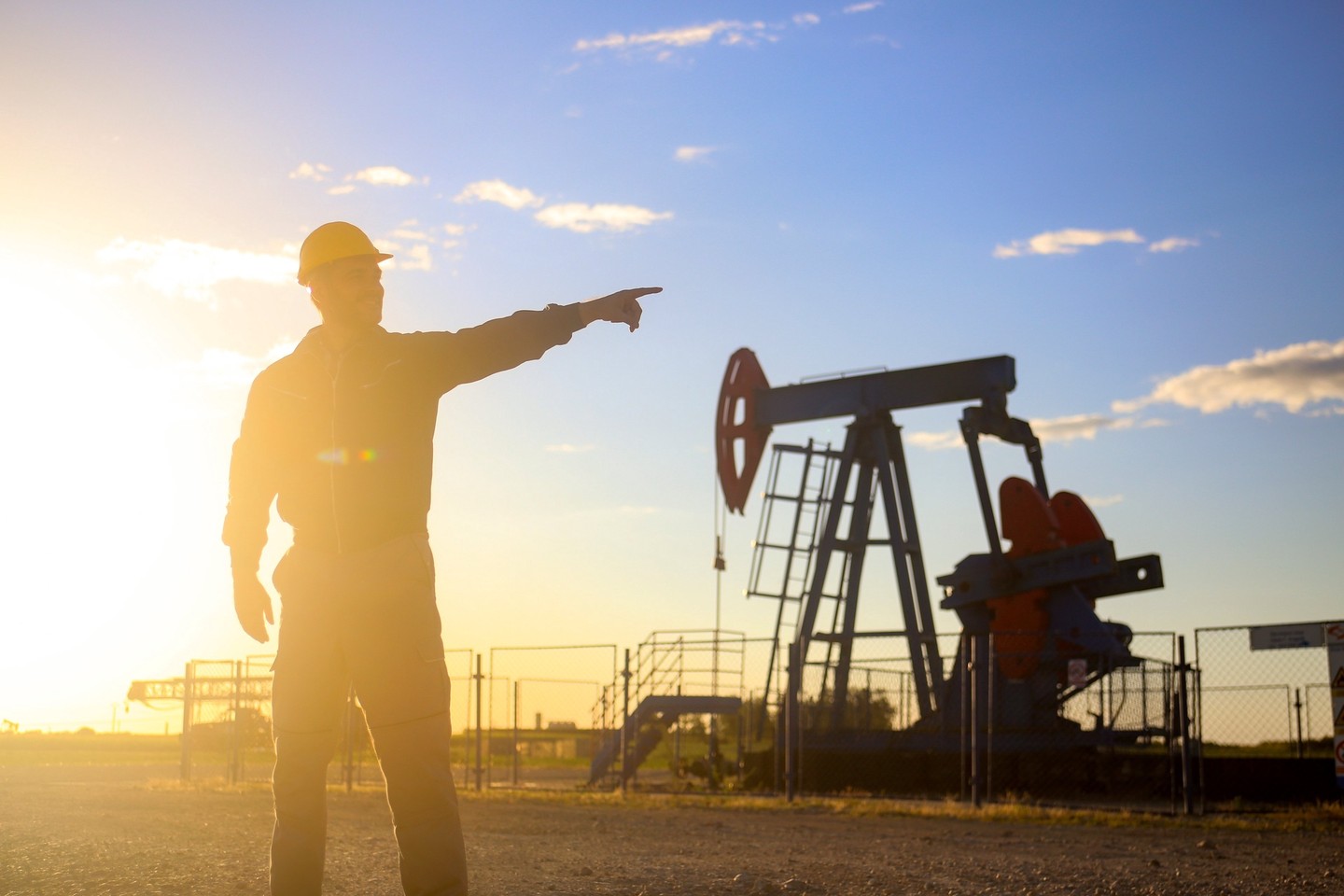 An oilfield worker points to a pumpjack against the backdrop of a setting sun on the horizon.